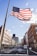 An American flag is prominently displayed, billowing in the wind against a clear, sunny sky. The scene features an urban street flanked by historic and modern buildings. Cars are parked along the street, and a few signs and pedestrians are visible. The atmosphere feels open and patriotic.