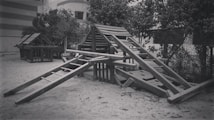 A monochrome photograph of a playground featuring wooden climbing structures and slides, set on a sandy ground. Trees and a building with striped walls are visible in the background, providing a natural and urban contrast.
