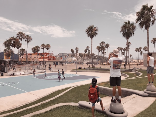 A scenic view of an outdoor basketball court surrounded by palm trees near a beach. People are either playing basketball or observing the game. Several colorful buildings and a clear sky are visible in the background, creating a relaxed, summery atmosphere.
