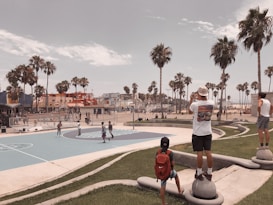 A scenic view of an outdoor basketball court surrounded by palm trees near a beach. People are either playing basketball or observing the game. Several colorful buildings and a clear sky are visible in the background, creating a relaxed, summery atmosphere.