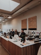 A modern coffee shop interior with three people standing behind the counter. The counter is made of wood with a white countertop, and various items such as containers, a coffee machine, and menu brochures are placed on it. The wall behind the counter features wooden menu boards listing different types of beverages. The establishment has a bright atmosphere with natural light coming from skylights on the ceiling and large windows on the left side.
