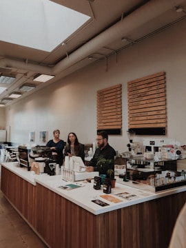 A modern coffee shop interior with three people standing behind the counter. The counter is made of wood with a white countertop, and various items such as containers, a coffee machine, and menu brochures are placed on it. The wall behind the counter features wooden menu boards listing different types of beverages. The establishment has a bright atmosphere with natural light coming from skylights on the ceiling and large windows on the left side.