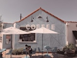 A quaint coffee shop with a light blue exterior and red tile roof. The word 'COFFEE' is painted in large white letters on the wall. Two white umbrellas provide shade over a few blue tables and chairs in the outdoor seating area. Two children sit at one of the tables.