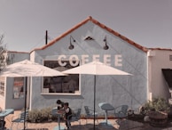 A quaint coffee shop with a light blue exterior and red tile roof. The word 'COFFEE' is painted in large white letters on the wall. Two white umbrellas provide shade over a few blue tables and chairs in the outdoor seating area. Two children sit at one of the tables.