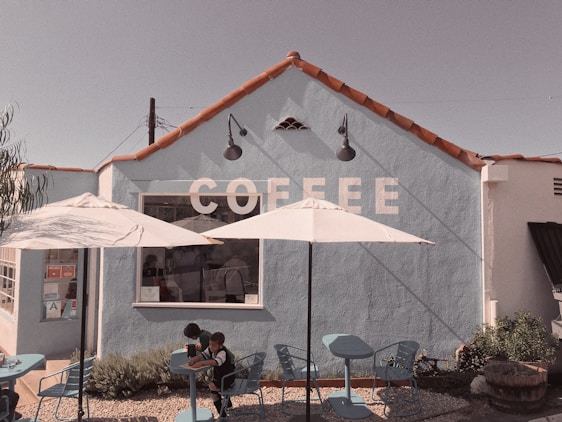 A quaint coffee shop with a light blue exterior and red tile roof. The word 'COFFEE' is painted in large white letters on the wall. Two white umbrellas provide shade over a few blue tables and chairs in the outdoor seating area. Two children sit at one of the tables.