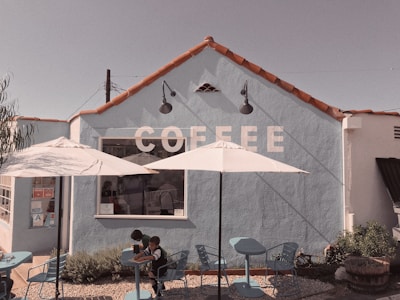 A quaint coffee shop with a light blue exterior and red tile roof. The word 'COFFEE' is painted in large white letters on the wall. Two white umbrellas provide shade over a few blue tables and chairs in the outdoor seating area. Two children sit at one of the tables.