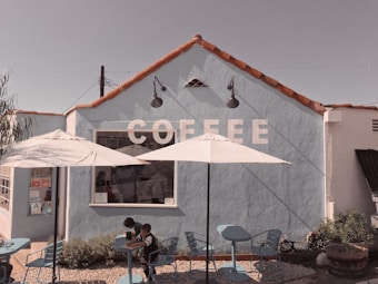 A quaint coffee shop with a light blue exterior and red tile roof. The word 'COFFEE' is painted in large white letters on the wall. Two white umbrellas provide shade over a few blue tables and chairs in the outdoor seating area. Two children sit at one of the tables.