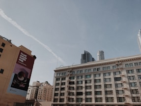 A cityscape featuring multiple buildings under a clear blue sky. A large rectangular billboard on one of the buildings reads 'The world needs your boundless energy' along with a logo. The scene includes a mix of older and modern skyscrapers, showing fire escapes and contrasting architectural styles.