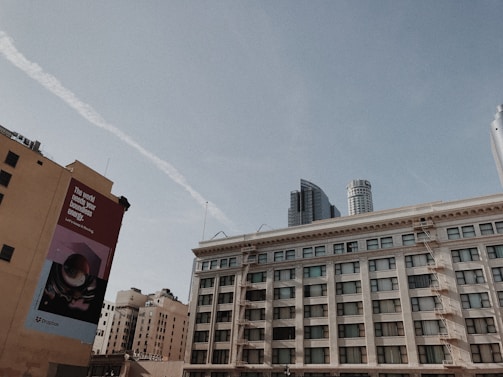 A cityscape featuring multiple buildings under a clear blue sky. A large rectangular billboard on one of the buildings reads 'The world needs your boundless energy' along with a logo. The scene includes a mix of older and modern skyscrapers, showing fire escapes and contrasting architectural styles.