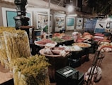 A market stall displaying a variety of fresh produce, including yellow asparagus, red chili peppers, and other vegetables. Baskets and crates are filled with different items, and the backdrop consists of informational signs and a soft overhead light.
