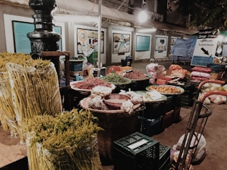 A market stall displaying a variety of fresh produce, including yellow asparagus, red chili peppers, and other vegetables. Baskets and crates are filled with different items, and the backdrop consists of informational signs and a soft overhead light.