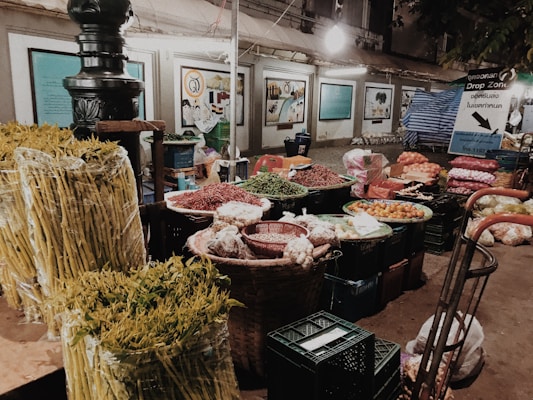 A market stall displaying a variety of fresh produce, including yellow asparagus, red chili peppers, and other vegetables. Baskets and crates are filled with different items, and the backdrop consists of informational signs and a soft overhead light.