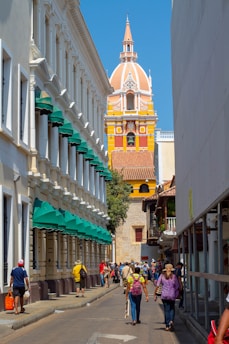 A vibrant street scene featuring a colorful historic church tower with intricate architecture as the focal point in the background. The street is lined with buildings having white facades and green awnings on one side, and more traditional structures on the other side. People are casually walking, some carrying bags or wearing hats, suggesting a tourist-friendly area with a lively atmosphere.