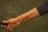 An arm is extended with a tattoo that features a heartbeat line, a red heart symbol, and the word 'Mom'. The person is holding a black object in their hand, possibly a stick or a handle. The background is grassy and slightly out of focus.