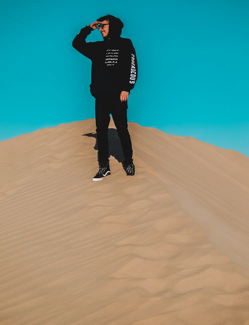 man wearing black pullover hoodie, black pants and black Vans OldSkool sneakers standing on desert sand