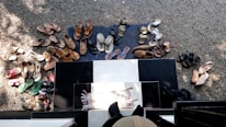 Numerous pairs of shoes are arranged on a gravel surface and a black and white tiled area at the entrance of a building. The shoes include a variety of sandals, slippers, and flip-flops in different colors and styles, indicating perhaps a social gathering or household activity. A small woven mat is placed at the door, and shadows fall across the scene, suggesting daylight.