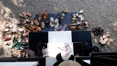 Numerous pairs of shoes are arranged on a gravel surface and a black and white tiled area at the entrance of a building. The shoes include a variety of sandals, slippers, and flip-flops in different colors and styles, indicating perhaps a social gathering or household activity. A small woven mat is placed at the door, and shadows fall across the scene, suggesting daylight.