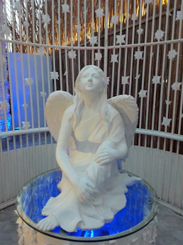 A white marble statue of an angel with flowing hair and wings sits on a glass surface. The angel gazes upwards with a serene expression. The background features a circular enclosure with white star decorations.