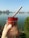 A close-up of a hand holding a plastic cup filled with a frozen dessert, which is dark red in color and has a wooden stick inserted into it. The background features a calm body of water, a clear blue sky, and some distant trees and buildings.