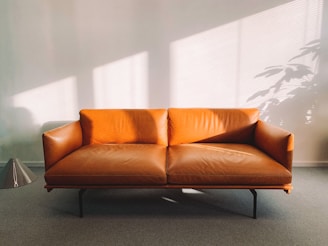 A stylish, modern brown leather sofa with sleek, angular design sits in a well-lit room. The light creates soft, elongated shadows on the wall, alongside a potted plant's silhouette. A metallic floor lamp is placed beside the sofa, contributing to a minimalistic aesthetic.