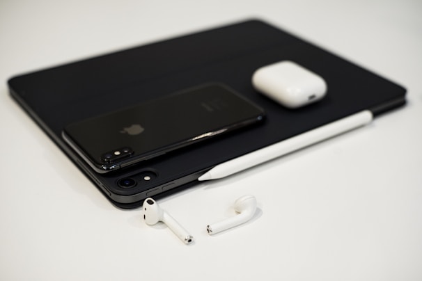 A collection of sleek electronic devices including a smartphone, tablet, stylus, wireless earbuds, and a charging case arranged on a white surface. The minimalist design and modern technology are accentuated by a monochrome color palette.