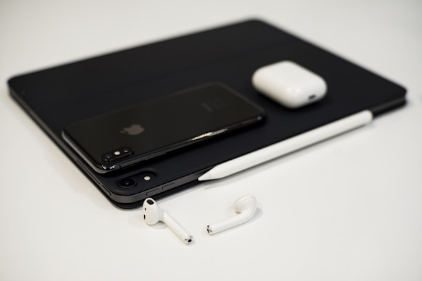A collection of sleek electronic devices including a smartphone, tablet, stylus, wireless earbuds, and a charging case arranged on a white surface. The minimalist design and modern technology are accentuated by a monochrome color palette.