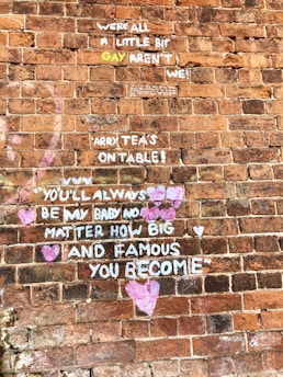 A brick wall features multiple graffiti messages in white and colorful paint. Phrases express sentiments of love, acceptance, and support, with several hearts drawn around the words. The background consists of reddish-brown bricks giving a rustic texture.