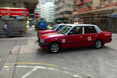 Two red taxis are in motion on a city street surrounded by blurred buildings and infrastructure, indicating a sense of speed and urban hustle. The taxis are prominently featured, with visible signage and a classic boxy design.