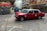 Two red taxis are in motion on a city street surrounded by blurred buildings and infrastructure, indicating a sense of speed and urban hustle. The taxis are prominently featured, with visible signage and a classic boxy design.