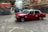 Two red taxis are in motion on a city street surrounded by blurred buildings and infrastructure, indicating a sense of speed and urban hustle. The taxis are prominently featured, with visible signage and a classic boxy design.
