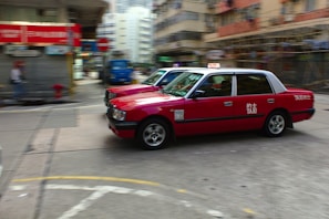 Two red taxis are in motion on a city street surrounded by blurred buildings and infrastructure, indicating a sense of speed and urban hustle. The taxis are prominently featured, with visible signage and a classic boxy design.