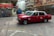 Two red taxis are in motion on a city street surrounded by blurred buildings and infrastructure, indicating a sense of speed and urban hustle. The taxis are prominently featured, with visible signage and a classic boxy design.