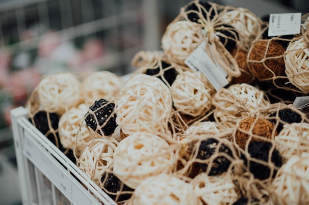 A collection of decorative rattan balls in various shades of cream, black, and brown, all enclosed in netting and placed in a white display basket with price tags attached.