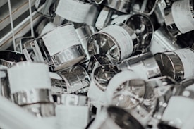 A pile of stainless steel flour sifters, some with visible labels, scattered in a metal bin. The image features an assortment of these kitchen tools, characterized by gleaming metallic surfaces and wire mesh interiors.
