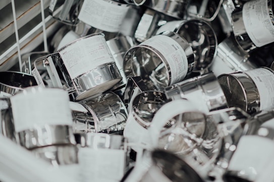 A pile of stainless steel flour sifters, some with visible labels, scattered in a metal bin. The image features an assortment of these kitchen tools, characterized by gleaming metallic surfaces and wire mesh interiors.