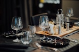 A dining table set elegantly with black plates, wine glasses, and cutlery. A wooden tray holds napkins, salt and pepper shakers, and possibly a small teapot or pitcher. The atmosphere suggests a fine dining setting with dim lighting.