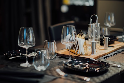 A dining table set elegantly with black plates, wine glasses, and cutlery. A wooden tray holds napkins, salt and pepper shakers, and possibly a small teapot or pitcher. The atmosphere suggests a fine dining setting with dim lighting.
