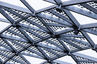 An intricate geometric structure featuring metal beams arranged in a radial pattern. The beams are intersected by square grid patterns, lending a modern and industrial aesthetic.