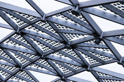 An intricate geometric structure featuring metal beams arranged in a radial pattern. The beams are intersected by square grid patterns, lending a modern and industrial aesthetic.