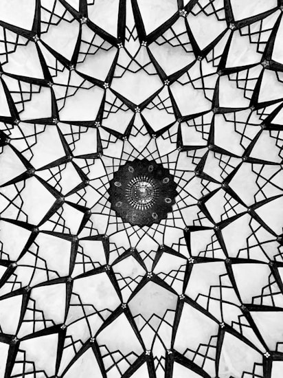 An intricate geometric pattern resembling Islamic art, featuring a series of interconnected shapes radiating from a central point. The design primarily uses a monochrome color palette, giving it an elegant and timeless quality.