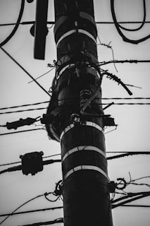 A black and white image of an electrical pole. Numerous cables and wires are wrapped around and connected to the pole, with various insulators and connectors visible. The background is a clear or overcast sky, contributing to a stark contrast.