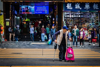 An elderly person with a crutch crosses the street while carrying a brightly colored rolling suitcase. A crowded area with people walking in various directions, some browsing or entering shops. Brightly lit electronic signs and numerous storefronts are visible, suggesting a busy urban setting.