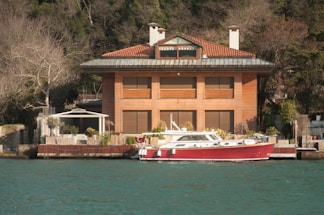 A large, modern, three-story house with a red-tiled roof and wooden exterior is situated by the water. In front of the house, there is a dock with a red and white boat moored. The surrounding area is lush with trees, though some of the trees appear bare, suggesting a non-summer season. The water has a greenish-blue hue, and the sky is clear, indicating good weather.