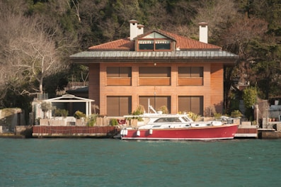 A large, modern, three-story house with a red-tiled roof and wooden exterior is situated by the water. In front of the house, there is a dock with a red and white boat moored. The surrounding area is lush with trees, though some of the trees appear bare, suggesting a non-summer season. The water has a greenish-blue hue, and the sky is clear, indicating good weather.