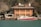 A large, modern, three-story house with a red-tiled roof and wooden exterior is situated by the water. In front of the house, there is a dock with a red and white boat moored. The surrounding area is lush with trees, though some of the trees appear bare, suggesting a non-summer season. The water has a greenish-blue hue, and the sky is clear, indicating good weather.