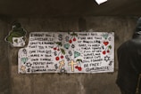A piece of street art on a concrete wall. The artwork features hand-drawn text and various symbols like crosses, dollar signs, and cherries. A cartoonish face with green hair appears on the left. The message in the text involves themes of self-identity and critiques on money and fear. A person in a dark hoodie stands to the right, partially obscured.