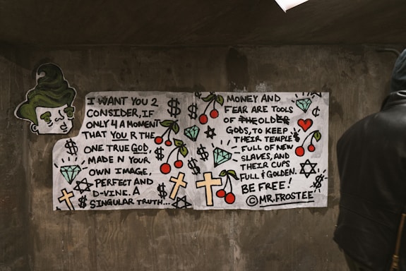 A piece of street art on a concrete wall. The artwork features hand-drawn text and various symbols like crosses, dollar signs, and cherries. A cartoonish face with green hair appears on the left. The message in the text involves themes of self-identity and critiques on money and fear. A person in a dark hoodie stands to the right, partially obscured.