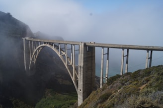 A large, arched concrete bridge spans across a deep valley surrounded by fog. The structure is set against a backdrop of steep hills and lush greenery. The scene conveys a sense of majesty and engineering prowess.