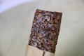 A close-up view of a brown, textured, rectangular granola bar partially wrapped in light brown paper.