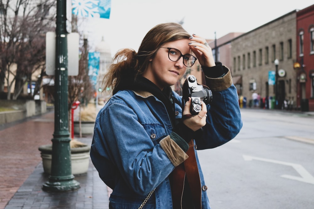 woman in blue denim jacket holding camera while standing on road side